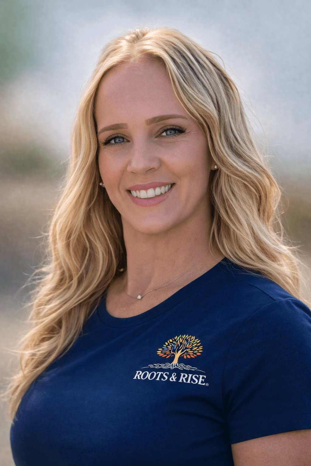 Smiling blonde woman wearing a navy blue shirt with a colorful Roots & Rise logo.